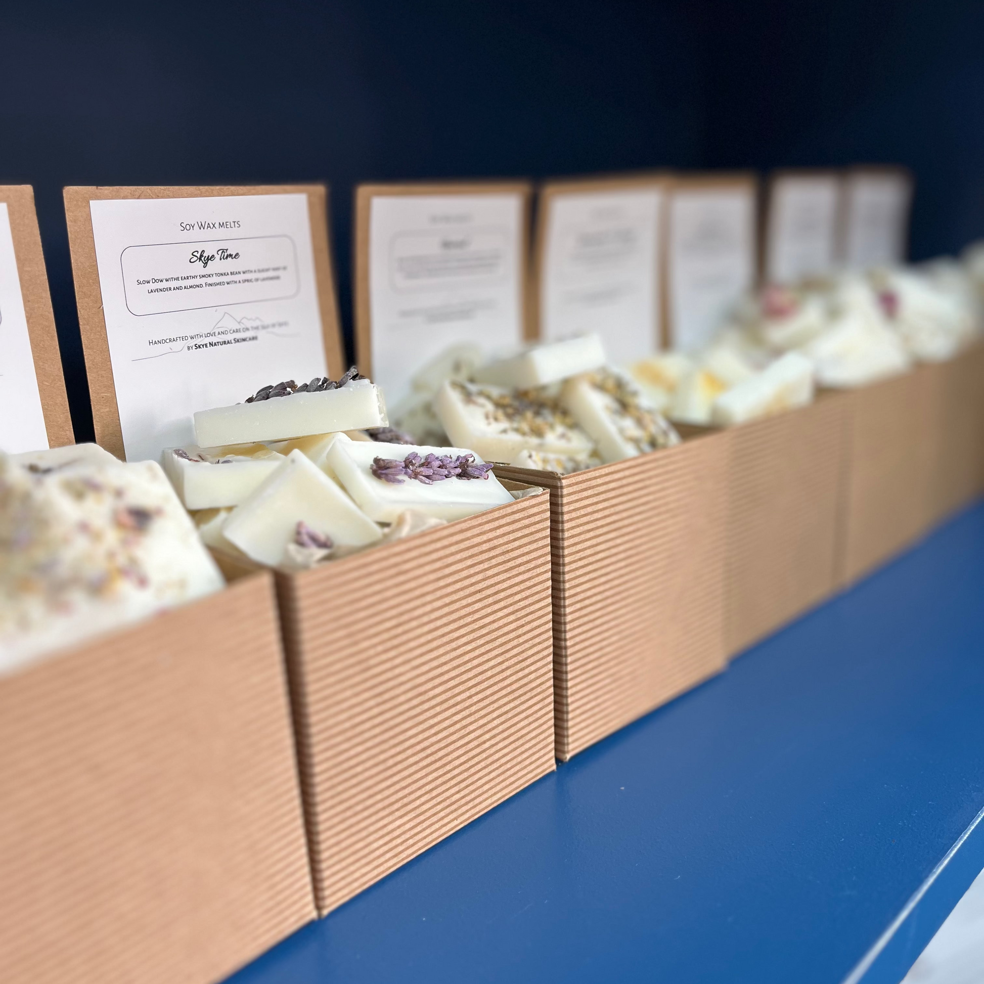 Row of wax melts in cardboard boxes with labels on a blue surface.