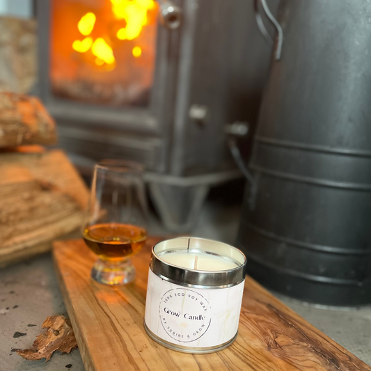 Candle in a tin with a glass of whiskey on a wooden board in front of a lit wood stove.